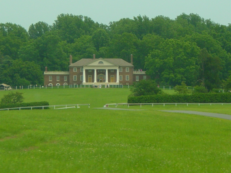 A brick home with white columns sits on a grassy hill with tall trees behind it