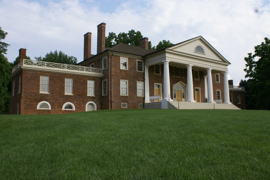 A brick home with white columns sits on a grassy hill with tall trees behind it