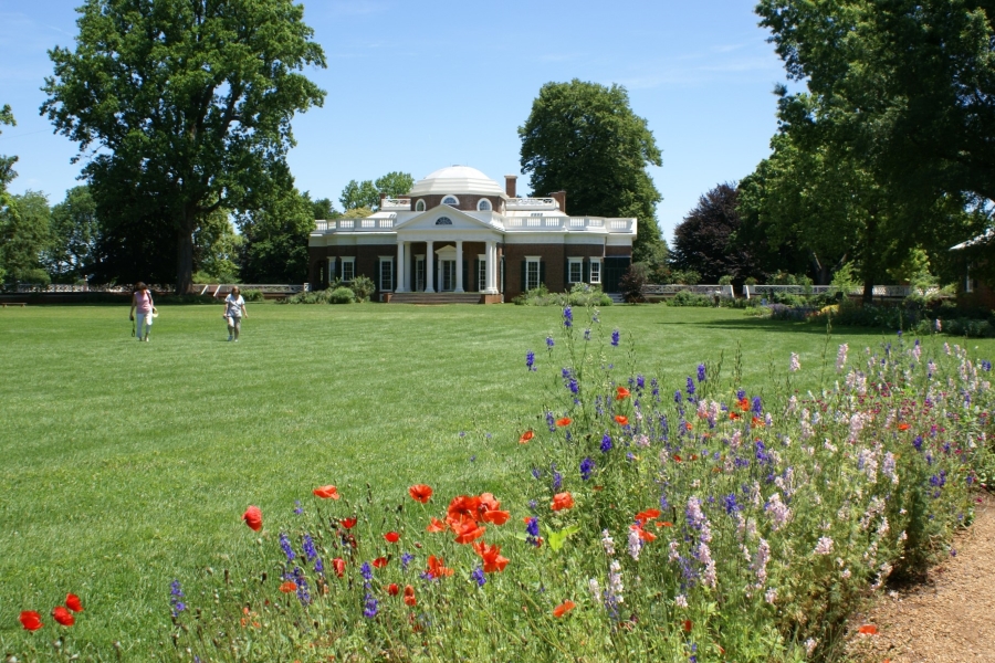 Flowers bloom along a path surrounding an open green grass lawn in front of the brick building with a stark white dome