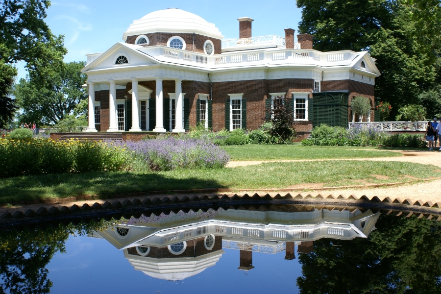 A brick building with a white dome over the center and white columns is reflected in the still water of a small pool