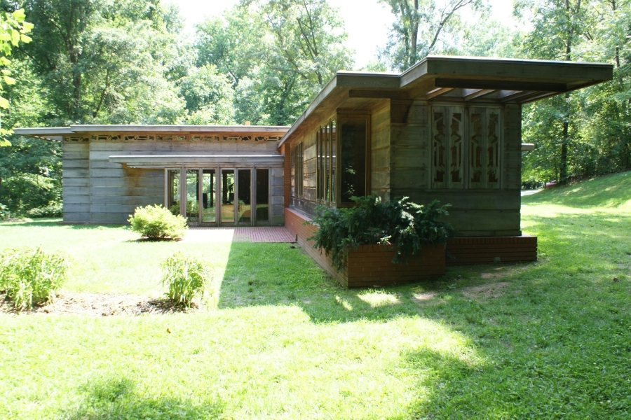 A “L” shaped building made of brick and wood with large windows below extending flat overhangs sits amongst large trees