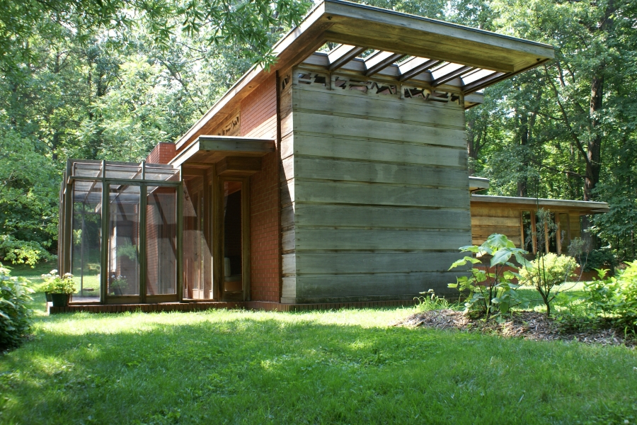 A wood framed structure extends beyond a faded wood paneled wall next to a solid brick wall with a small patio