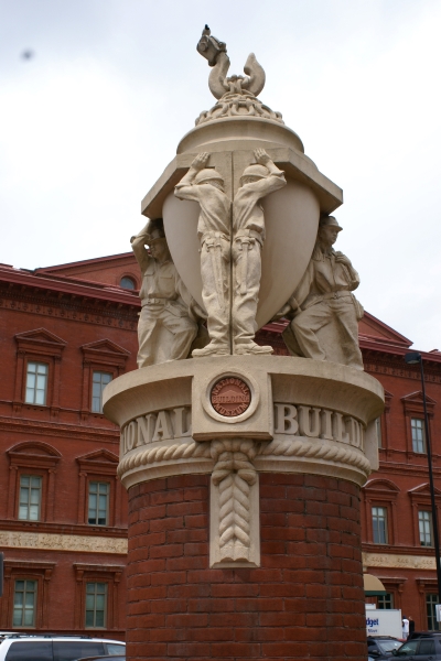 A large diameter round brick column supports stone sculpture of men supporting a dome