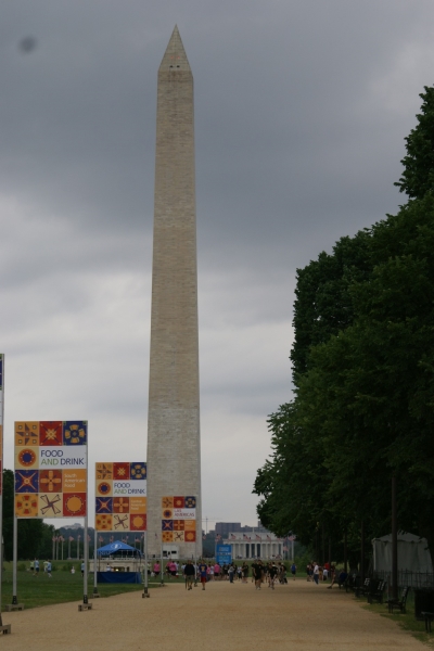 A thin white obelisk extends into a cloudy sky above a green park with a tan stone walking path