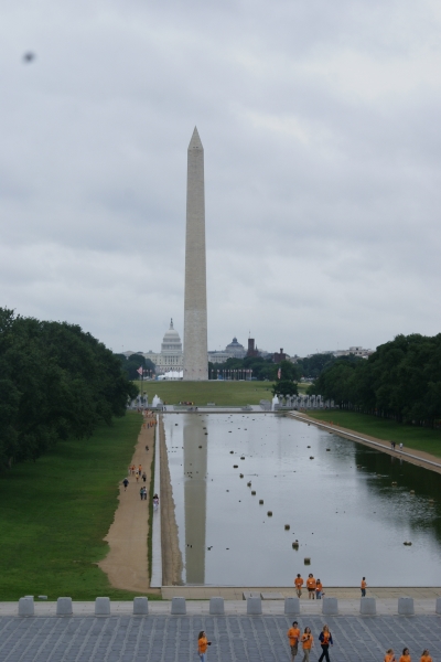 The Washington Monument is reflected in a rectangular pool of water with cloudy skies and the Capitol building beyond