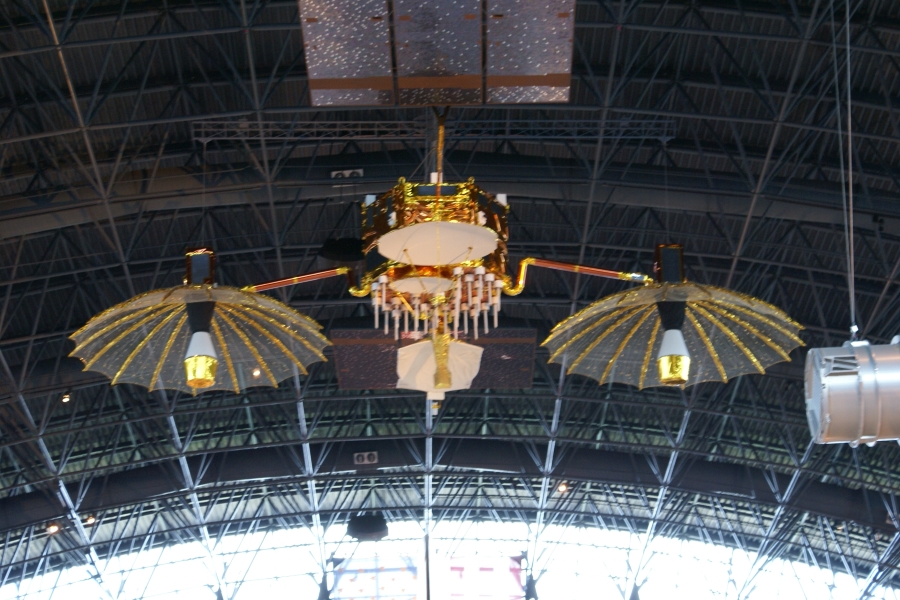 A satellite with two bright gold dishes hangs from the roof structure of a arched building