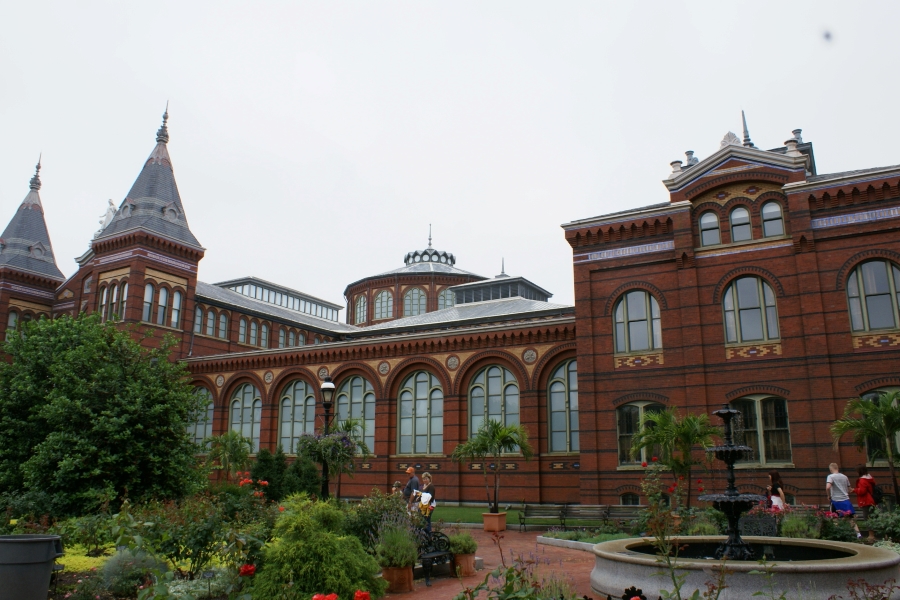 Gardens and paths run near a large red brick building with gridded arch top windows, cupolas near the entry and a large dome over the central portion of the building