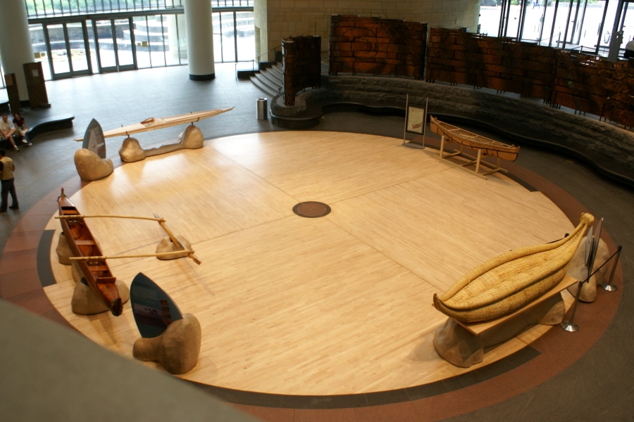 A wooden circle sits in an atrium space with four canoes made of various materials and in different styles at the edge