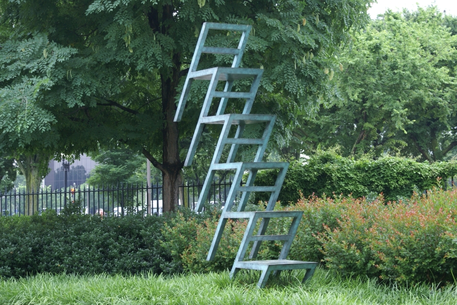 A sculpture of what appears to be stacked chairs with strangely angled elements stands on a small mound of green grass