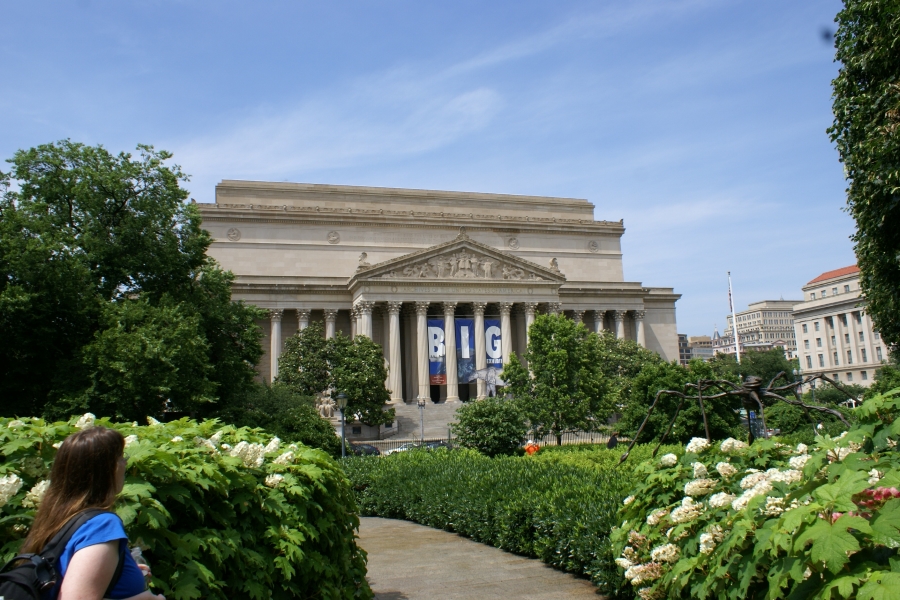 A walking path through green flowering bushes leads to a large white stone building with two-story columns and a triangular header over the entry