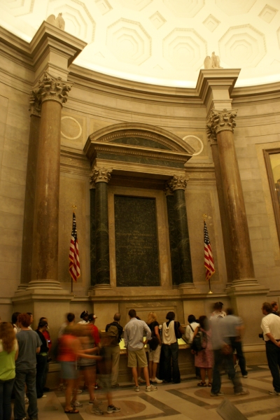 People stand in line to view a document in a protective case with marble columns with American Flags on each side