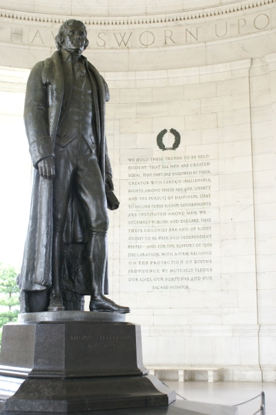 A large bronze statue of a man stands in the center of a circular space; the wall beyond has the open stanza of the Declaration of Independence carved in the white stone