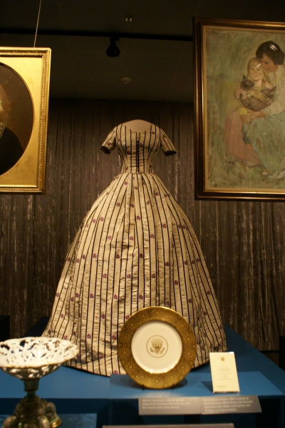 A dress with vertical striping, narrow waist, and voluminous petticoat worn by one of the early first ladies stands in a glass enclosed case