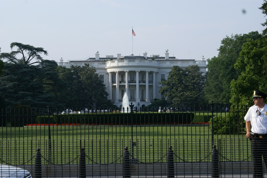A three-story white building with a curved portico created by columns sits amongst trees and green grass behind a black wrought iron fence