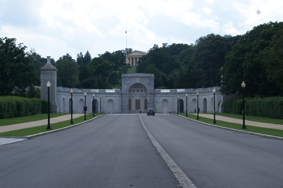 A curved wall of white stone with arches sits at the end of a wide boulevard in front of a green hill dotted with trees