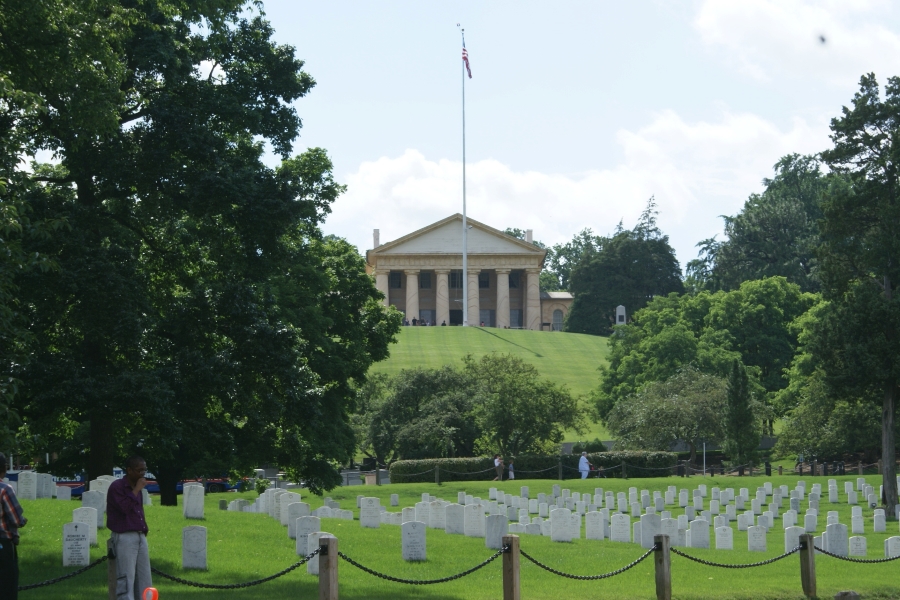 Rows of white stone markers fill a grassy field below a tan colonial building on a hill