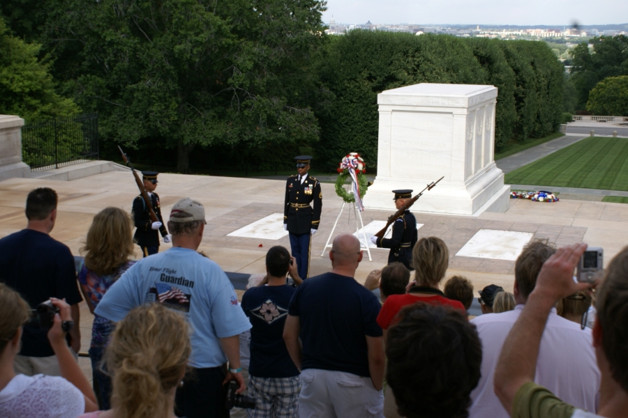 People watch a ceremony with three uniformed soldiers in front of a stark white crypt