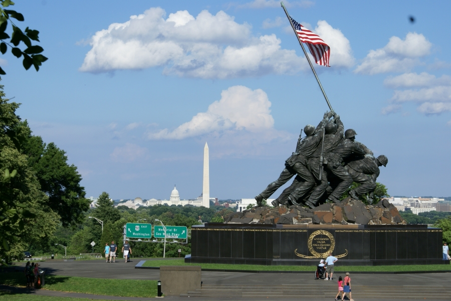 A very large statue of men working together to raise a flagpole stands in the road with monuments visible beyond