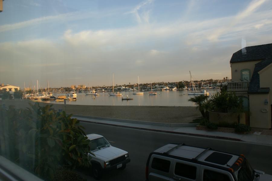 The yellowish light of dawn reveals scattered feathery clouds above sailboats anchored in a waterway between densely developed hillside and a small sandy beach between houses still in shadow