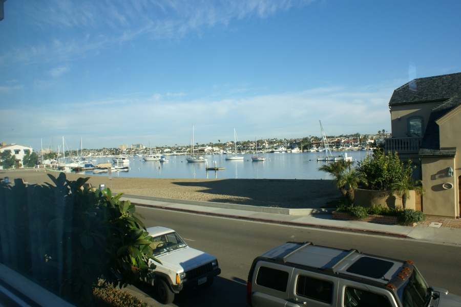 A bright blue sky with scattered feathery clouds above sailboats anchored in a waterway between densely developed hillside and a small sandy beach between houses across the street from the photographer