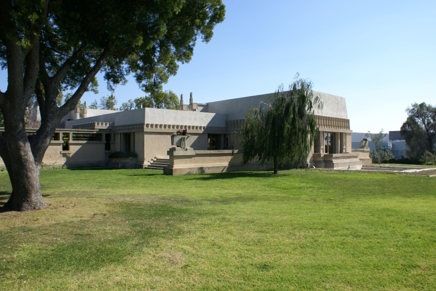 A green lawn with sparse trees leads to a low-profile tan stucco building with a small pool to one side and steps leading to an entrance behind a short screen wall