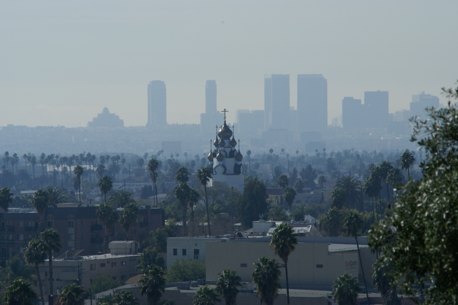 A church, with several onion shaped domes with crosses atop each, stands above the palm trees with high-rise buildings shrouded in yellowish smog beyond