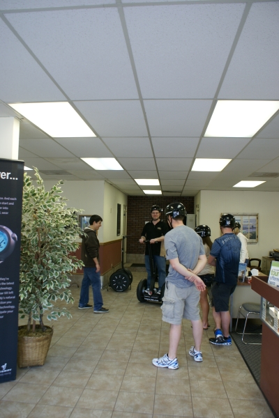 People look on in a shop as James learns how to control the movement of a two-wheeled device where the wheels are on the outside of his feet and a mast for his hands extends up for steering