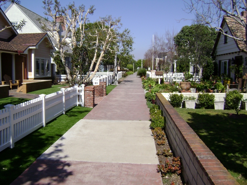 A narrow concrete and brick path runs between manicured lawns beyond short fences in a quite neighborhood