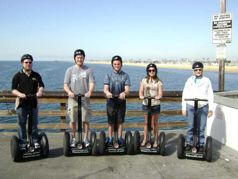 Five people stand on segways on a wooden pier with the ocean and a beach extending into the distance behind them