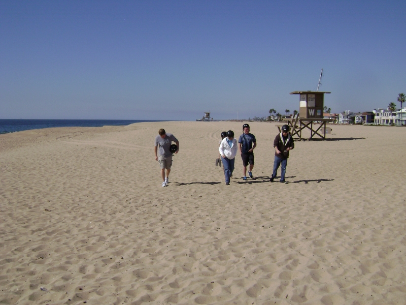 Five people walk through tan sand with the ocean to the left and a lifeguard building just behind them to the right