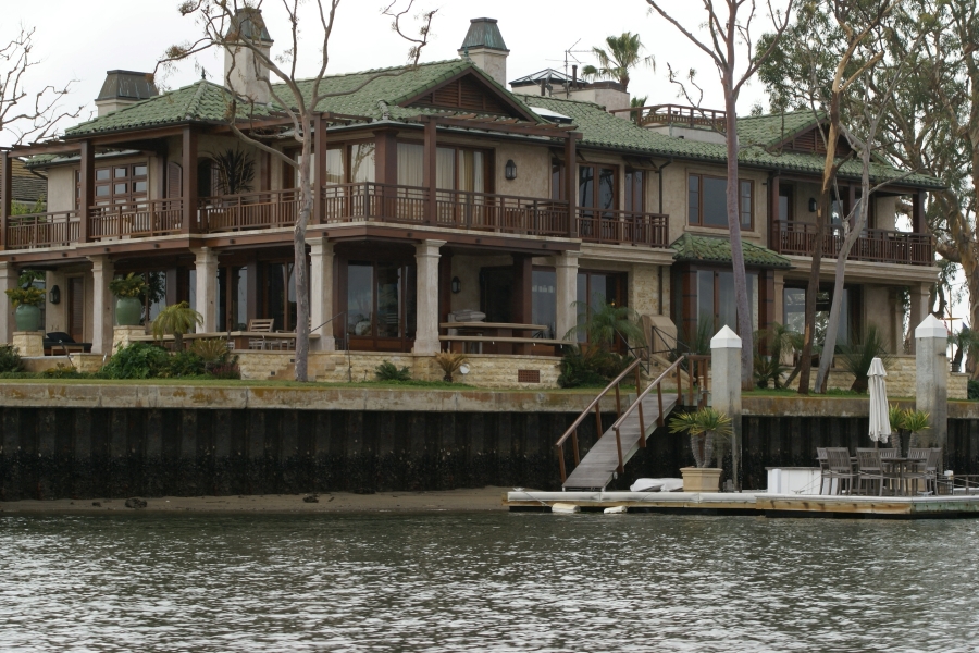 A large building with green slate roofs, tan walls and large windows behind porches covered with brown railed balconies sits amongst trees on a seawall with a ramp to a floating dock with picnic tables and chairs