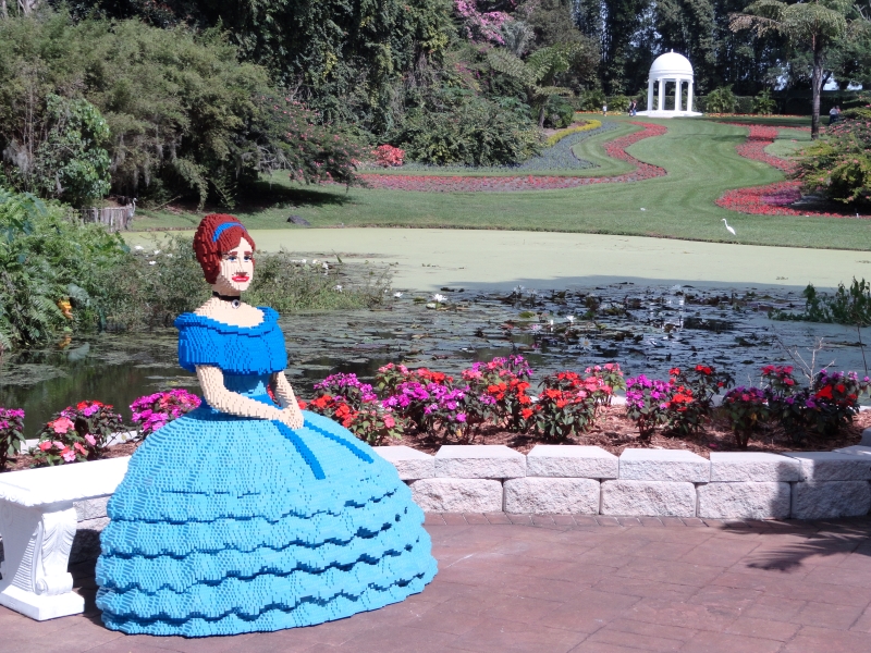 A life-size LEGO figure of a woman in an antebellum style blue dress sites on a bench near a pond at the base of a hill with a manicured lawn and small domed structure at the top