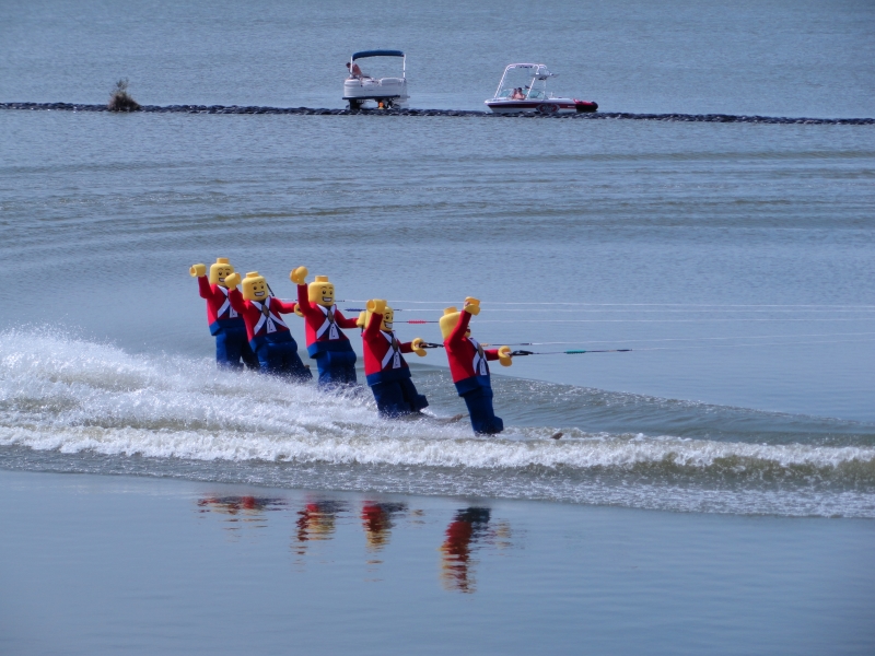 Five people dressed like LEGO minifigures with blue pants and red coats on waterskies slide across the water in formation