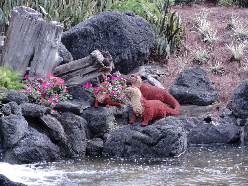 Three brown otters made of LEGO stand amongst real rocks and branches next to a waterfall