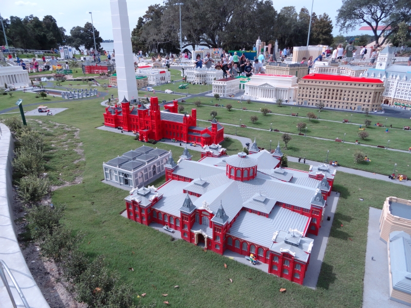 LEGO Models of red brick buildings, a large white obelisk, and other structures surround a grassy area with walkways and small people made from LEGO pieces