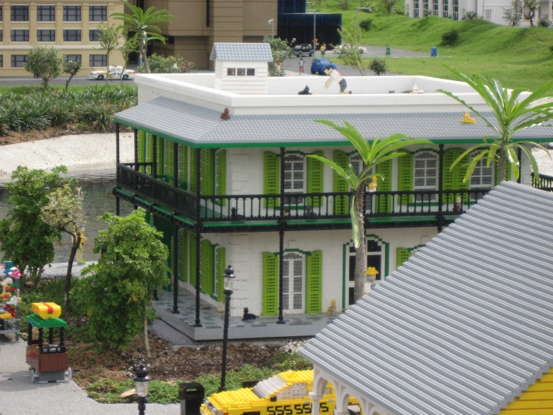 A square building with white walls and green shuttered windows with LEGO cats all over its roof, covered balconies and patios