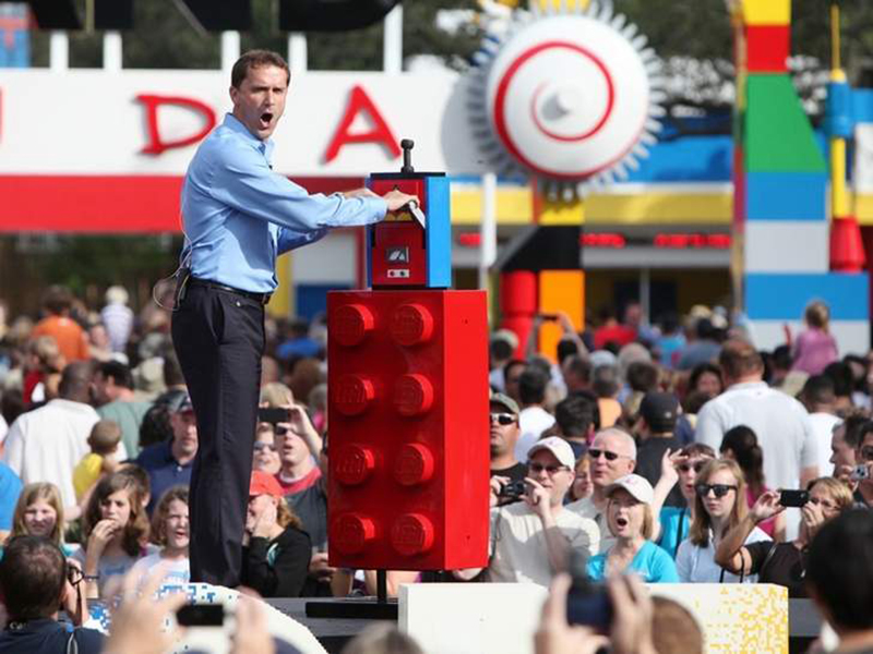 A man in a suit stands on a stage surrounded by croweds next to a LEGO shaped pstand with a switch and gates to a theme park in the distance