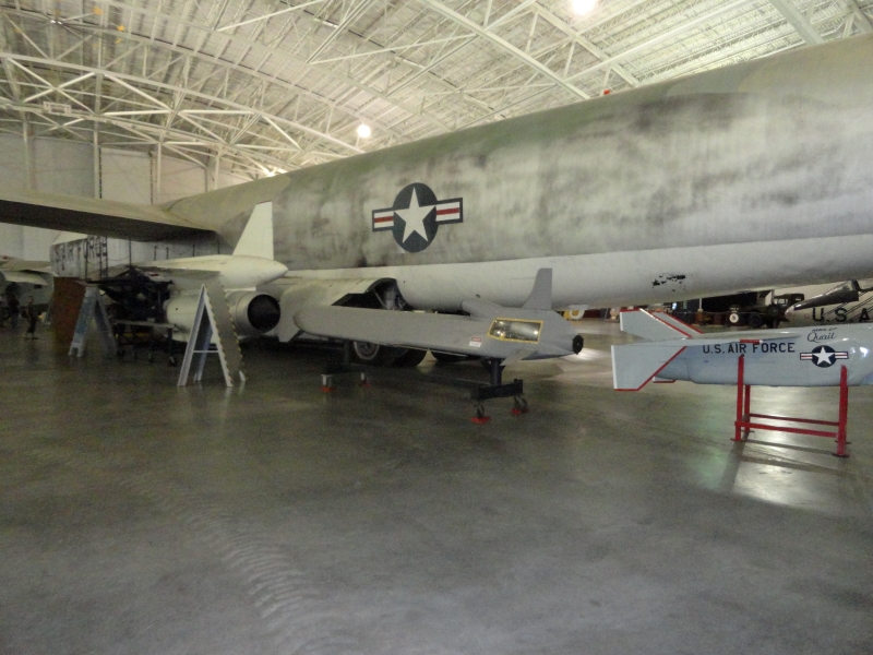 A small cruise missile with fins and an exhaust port sits on a rack near a large silver plane