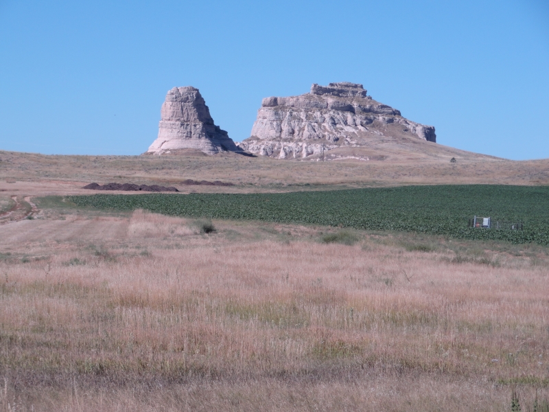 Gray stone buttes extend into the blue sky above fields of grass and corn