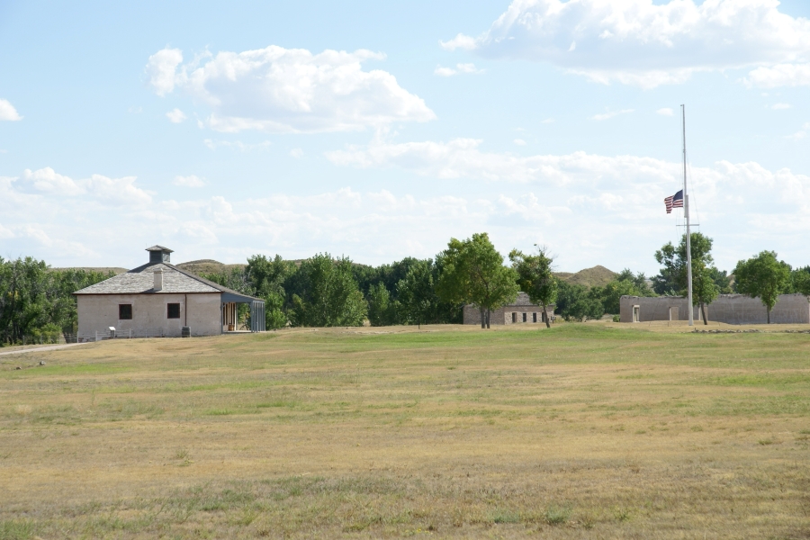 A flag flies at half-staff near remnants of old stone structures in a grassy plain surrounded by trees