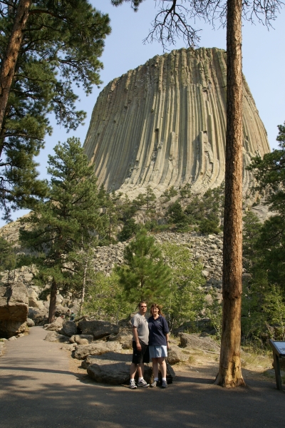 Julie and James stand amongst sparse trees along a walking path with a stone tower sticking up from a stone rubble field beyond