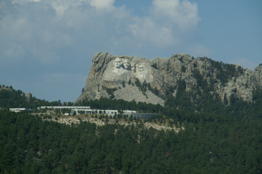 A stone hill stands above a green forested valley and a visitor center with the faces of four presidents carved into the cliff face