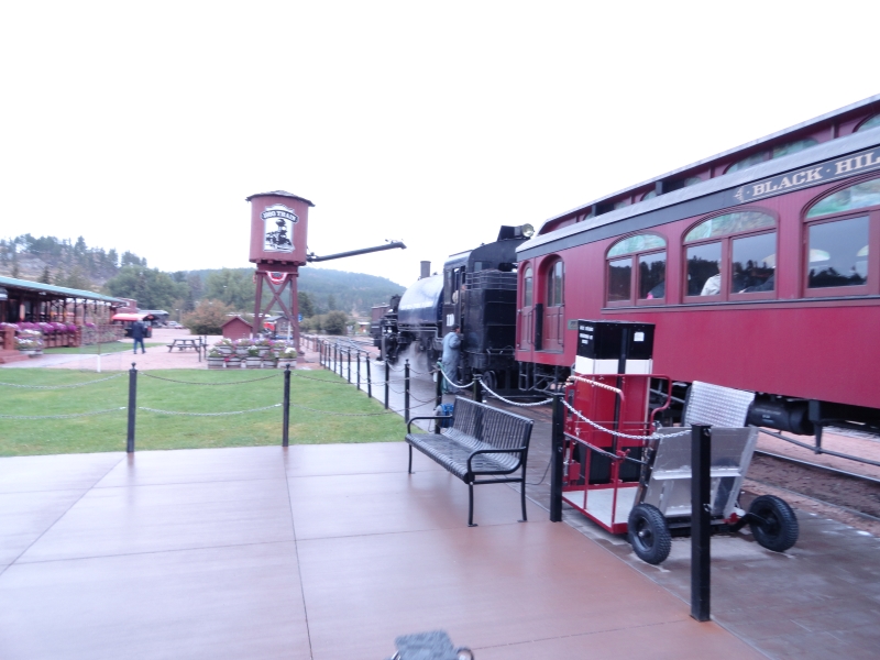 A water tower stands with a boom extended over a train line where a black steam engine sits in front of a passenger car painted marron and labeled Black Hills Express