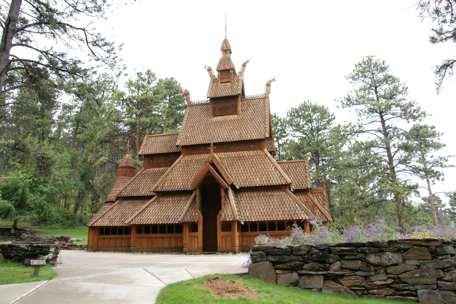 A wooden structure with steep stepped wood shingle roofs and carved decorations at the corners sits surrounded by tall pine trees and a low stone wall