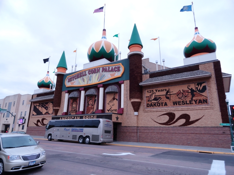 A building with ornate onion domes and rounded tower like elements covered in pictures made of colorful fibrous materials stands along the street with a bus parked in front