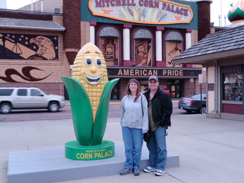 Julie and James stand next to a brightly painted statue of an ear of corn with the Mitchell Corn Palace across the street in the background