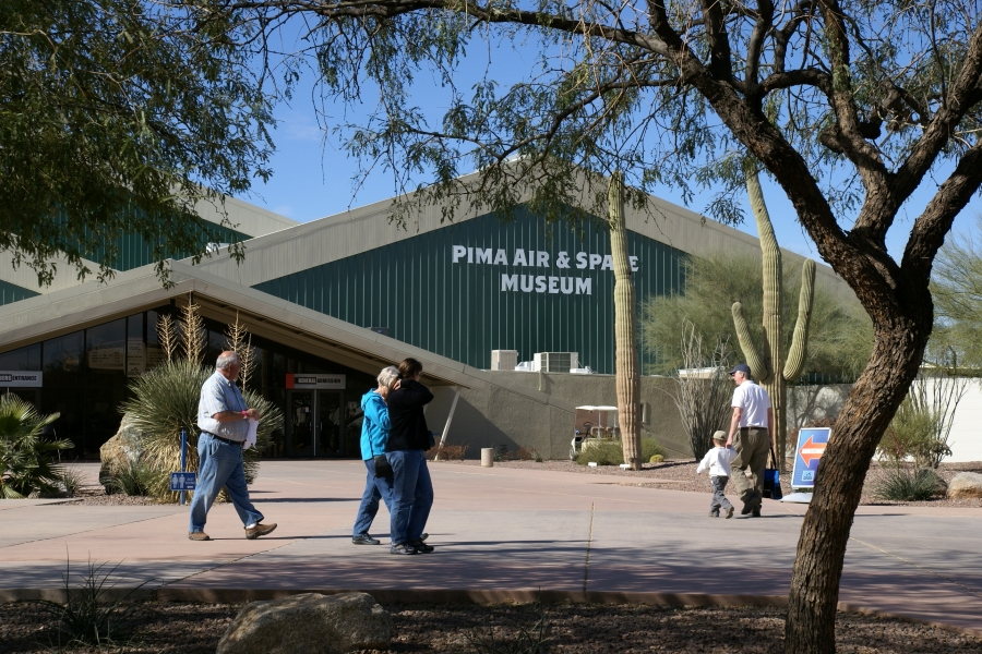 Julie walks with her parents under a canopy of scrubby trees near the entrance to a green building with diamond shape features