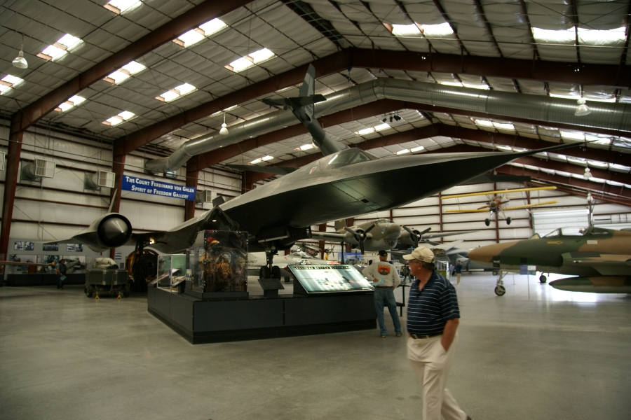 A black plane with unique wedge-shaped body and a pointed nose sits inside a large hanger with other aircraft