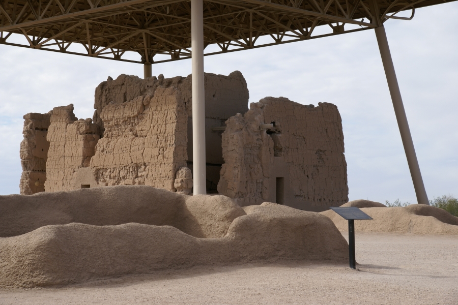 Worn adobe walls about 3-foot high with rounded tops extend above the soil with the walls of the great house beyond