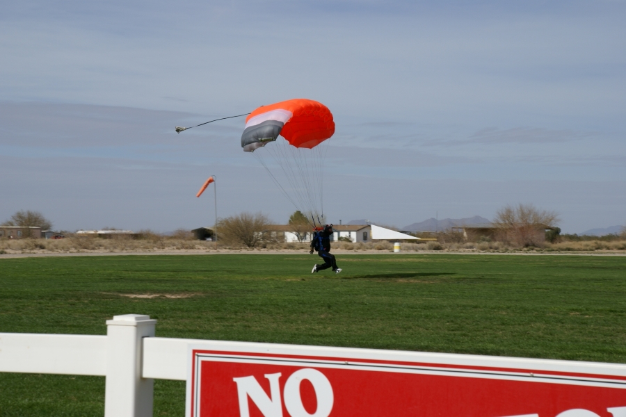 A person in a black suit drags a foot on an area of green turf as the touch down with their orange and gray parachute extends above their head